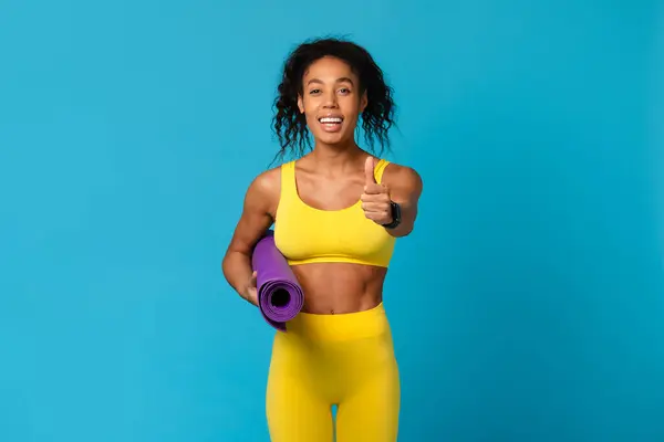 青い背景に対して黄色いスポーツ服に親指を与える紫色のヨガマットを保持している笑顔のアフリカ系アメリカ人女性 — ストック写真