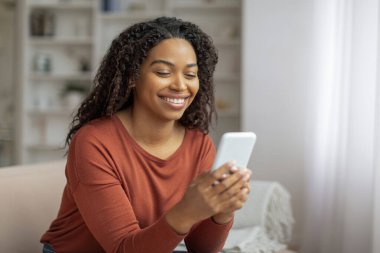 Gülümseyen siyah kadın bir koltukta oturuyor, elinde cep telefonu ekranına odaklanmış, Afro-Amerikan kadın akıllı telefonuyla evde mesajlaşıyor.