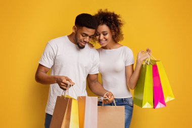 Gülümseyen genç Afro-Amerikan çift, parlak sarı bir arka planda neşe dolu alışveriş torbalarına bakıyorlar. Başarılı bir alışveriş çılgınlığı öneriyorlar.