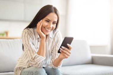 Mutlu bir gülümsemesi olan bir kadın akıllı telefona bakıyor, parlak ve havalı bir oturma odası kanepesinde oturuyor, boşluğu kopyalıyor