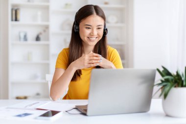 Dost canlısı gülümseyen, kulaklık takan ve bilgisayarından biriyle konuşan Asyalı genç bir kadın.