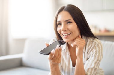 A joyful woman is seen holding a smartphone near her mouth, presumably using voice control or a voice assistant feature, copy space clipart
