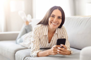 Neşeli bir kadın rahat bir şekilde koltuğa oturmuş, akıllı telefonuyla meşgul, mutlu bir tavır sergiliyor.
