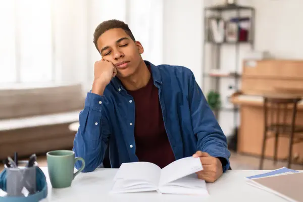 身穿蓝色衬衫的疲惫不堪的黑人年轻人把头靠在手上 一边在家里明亮的房间里从白书桌边看书 一边打瞌睡 — 图库照片