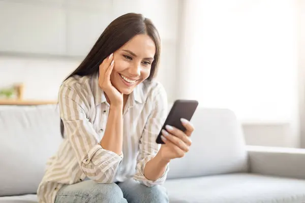 Mulher Com Sorriso Contente Olhando Para Smartphone Sentado Sofá Sala — Fotografia de Stock