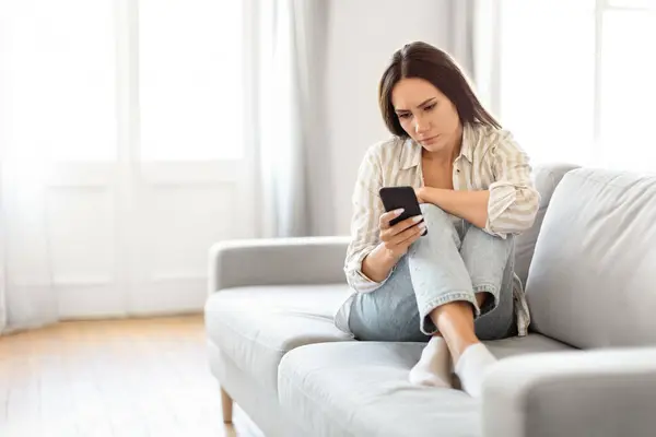 女性は自宅の明るい部屋でソファーに座ってスマートフォンを保持しているように ペンシルベニアで熟考しているように見えます — ストック写真
