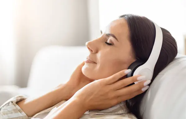 一个疲惫不堪的女人躺在沙发上 头戴耳机 带着科技的气息描绘着一种放松的生活 — 图库照片