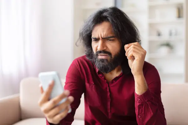 Indian man frowning and showing confusion while looking at his smartphone screen, got weird message
