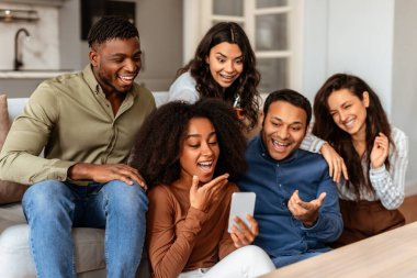Çok ırklı arkadaşlar, rahat bir ev ortamında akıllı telefona bakarak birbirlerine yakın bağlarını göstererek aynı kahkahayı atarlar.