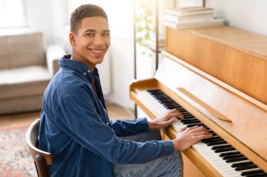 Mavi tişörtlü mutlu genç adam piyano başında oturuyor, kameraya bakarken gülümsüyor ve oyun oynarken gülümsüyor, ev ortamında neşeyle dolu.