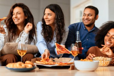 Çok ırklı arkadaşlar, pizza ve şarap dilimleriyle bir yaşam alanı etrafında toplanıyor. Sıcak bir atmosferde yemek ve sohbet paylaşıyorlar.
