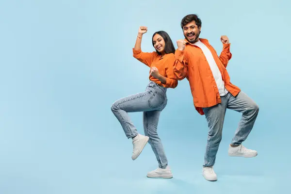 陽気な若いインドのカップルはオレンジのジャケットを着て デニムは青い背景に対して楽しいポーズで彼らの腕で踊っています — ストック写真