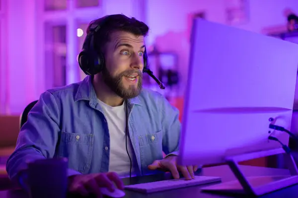 身着斜纹棉布衬衫的男人 手心紧握着 神魂颠倒 沉浸在电脑作业或游戏中 坐在霓虹灯照亮的桌子前 — 图库照片