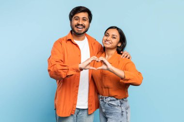 Gülümseyen Hintli çift elleri ile bir kalp şekli oluşturur, mavi arka planda aşkı ve bağlantıyı sembolize ederler.