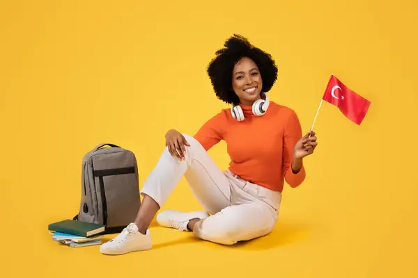 一个快乐的女人 脖子上戴着耳机 拿着土耳其国旗 背着书本和灰色背包 身穿舒适的橙色高领毛衣和白色裤子 背景是黄色的 — 图库照片