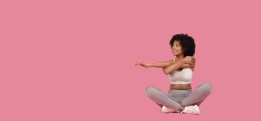 Smiling young african american woman in gray sportswear sits cross-legged, stretching her arms on a pink background clipart