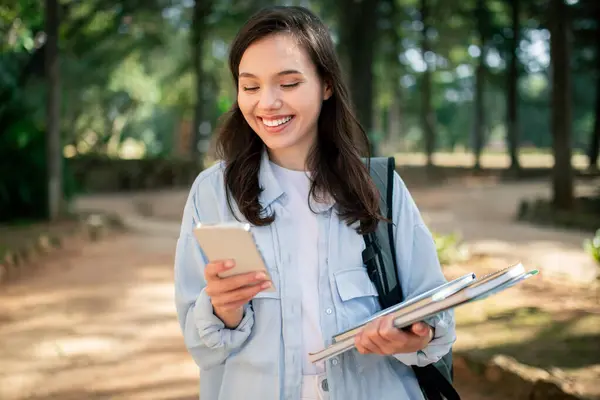 A young lady student engaged with smartphone and holding notebooks at public park, enjoying nice weather