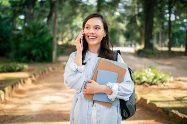 Neşeli genç bir kadın açık hava sahnesinde elinde defterlerle akıllı bir telefonla iletişim kuruyor.