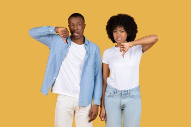 Afro-Amerikalı çift sarı arka planda onaylamadıklarını ve duraksadıklarını göstererek başparmaklarını indiriyor.