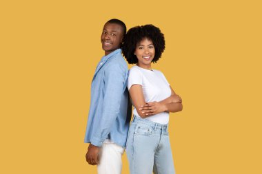 Two smiling young African American couple stand back to back, comfortably posing over a yellow background, exuding confidence and contentment clipart