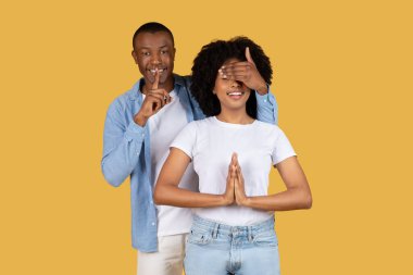 Umut dolu bir dilek dilerken Afro-Amerikalı bir adam bir kadının gözlerini kapatıyor. İkisi de sarı bir arka planın önünde duruyor.