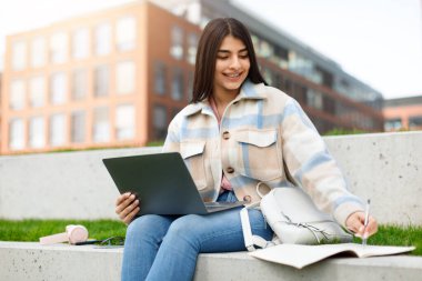 Sıradan bir ceket giymiş odaklanmış genç bir kız dışarıda bir bankta otururken laptopunu dikkatle kullanır.