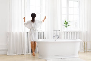 Beyaz cüppeli sakin bir Afro-Amerikalı kadın, aydınlık, havadar bir odada, pencerenin önünde duruyor, sakin ve lüks bir hissi uyandırıyor.