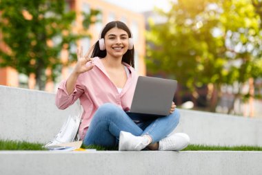 Laptop ve kulaklıklı mutlu kız dışarıda barış işareti yapıyor, odaklanmış ve neşeli bir ifadeyle.