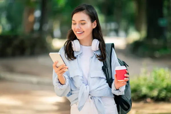 A young woman enjoys a walk in the park looking at her phone with headphones and a coffee, going home from college