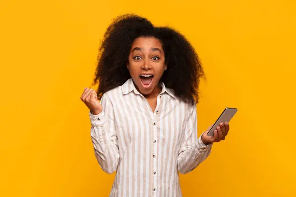 stock image Smiling young black lady thrilled with what she sees on her phone screen against yellow backdrop