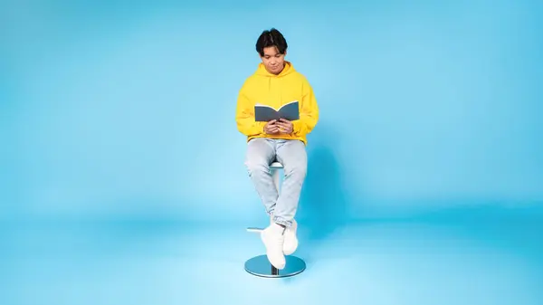 読書に巻き込まれて 若いアジアの男は青いスタジオの背景に対して本を持ってスツールの上に座ります — ストック写真