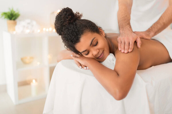 An image capturing a masseur providing a shoulder massage to a relaxed african american woman laying on a spa table
