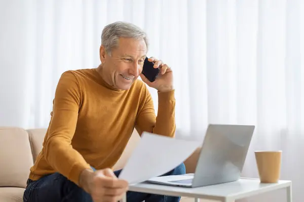 マスタードセーターの成熟した男は ラップトップを使用して 自宅で電話で話している間 慎重に紙を見ています — ストック写真