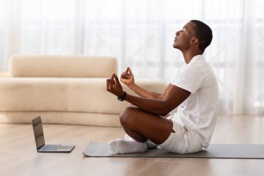Sakin bir yerde, laptopun önünde yoga minderi üzerinde meditasyon yapan, sağlığını ve farkındalığını arttıran odaklanmış genç bir zenci.