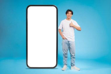Asyalı ergen bir adam mavi bir arka fona karşı dev bir akıllı telefon modeliyle baş parmak işareti yapıyor.