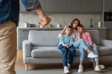 Görüntü gergin bir durumu tasvir ediyor. Bir anne, iki çocuğunu koltukta rahat ettirirken ve bir baba ön planda yumruğunu sıkarken.