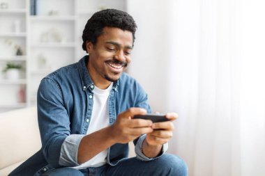 Yetişkin siyah bir erkek evde akıllı telefonuyla oyun oynuyor, cep telefonuyla eğleniyor, kanepede rahat bir şekilde oturuyor..