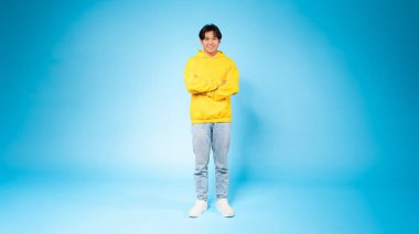 A cheerful young Asian guy stands confidently with arms crossed, wearing a bright yellow hoodie against a vivid blue background clipart