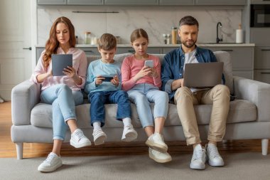 A family of four engrossed in digital devices while sitting together on a sofa in a cozy home setting clipart
