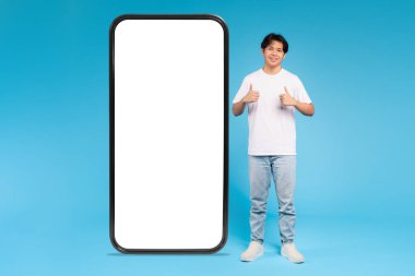 Neşeli bir Asyalı genç, büyük bir akıllı telefonun yanında duruyor. Reklam için boş bir ekranı var, mavi arka plan.