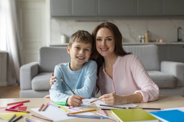 Gülümseyen bir anne, sanatsal eserlerini gururla sunarken oğlunu kucaklar. Masadaki renkli sanat malzemeleriyle çevrili olarak.