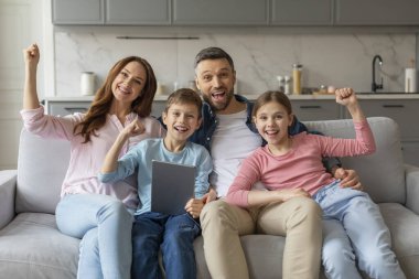 İki çocuklu gülümseyen bir aile kanepeye oturmuş, ellerinde dijital tabletle sevinçle yumruklarını kaldırıyorlar.