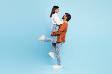 Neşeli bir Hintli adam kollarında bir kadını kaldırıyor, ikisi de mavi bir stüdyo arka planında günlük giysiler giyiyor, neşe ve mutluluk tasvir ediyor.