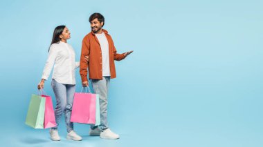 Alışveriş çantaları taşıyan Hintli çiftler birbirlerine şöyle bir bakıp mavi arka planda eğleniyorlar.