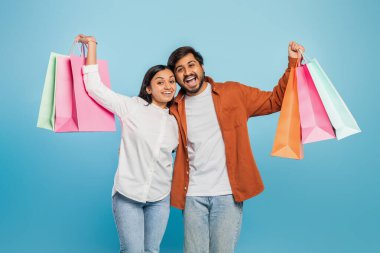 Genç bir Hintli kadın ve erkek, mavi bir arka planda birden fazla renkli alışveriş torbası taşıyarak mutlu olurlar.