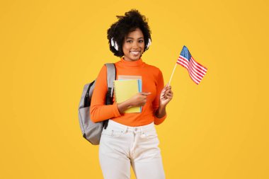 Amerikan bayrağı ve defterleri tutan, kulaklık takan ve sırt çantası takan, sarı arka planda, stüdyoda duran, gururlu Afrikalı Amerikalı bayan öğrenci. İngilizce kursları