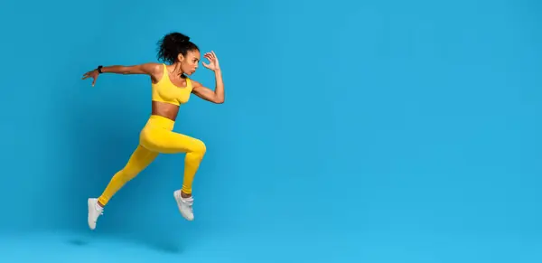 エネルギッシュな若いアフリカ系アメリカ人女性は 青い背景にダイナミックなミッドエアジャンプで撮影された黄色いスポーツ服を着ています — ストック写真