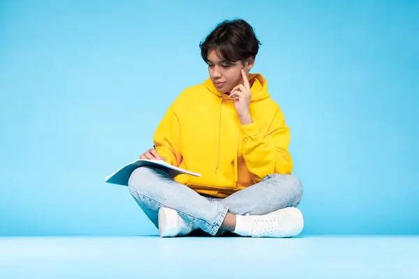 黄色いセーターの若いアジアのティーンエイジャーは 青い背景にオープンブックで横足に座っている間熟考 — ストック写真