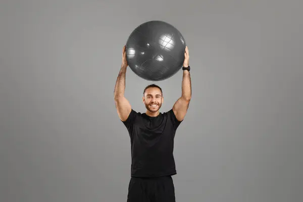 笑顔の男は 灰色の背景に強さと健康を示す彼の頭の上に大きなフィットネスボールを持ち上げます — ストック写真