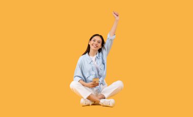 Cheerful young lady celebrating a success with a raised fist while sitting against an orange backdrop clipart